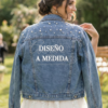 Novia sonriente en el jardín de su boda llevando una chaqueta vaquera personalizada con perlas y el texto "DISEÑO A MEDIDA" en la espalda, indicando servicio de diseño exclusivo