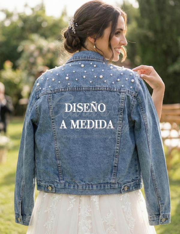 Novia sonriente en el jardín de su boda llevando una chaqueta vaquera personalizada con perlas y el texto "DISEÑO A MEDIDA" en la espalda, indicando servicio de diseño exclusivo