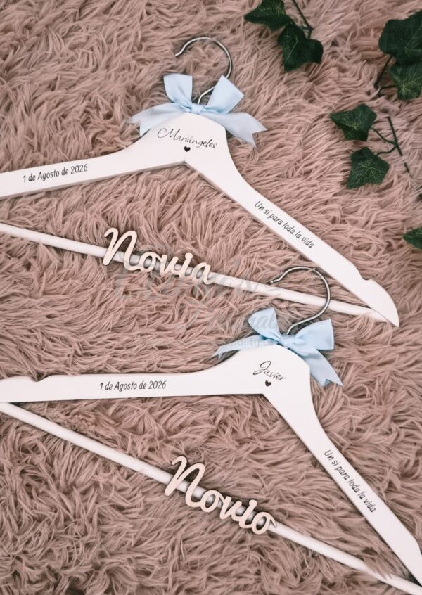 Perchas de madera personalizadas para novios en color blanco con nombres grabados Mariángeles y Javier, fecha de boda 1 de Agosto de 2026, lazos azul celeste y frase Un sí para toda la vida, creadas por Aura's Personality