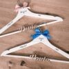 Pareja de perchas de madera personalizadas para novios en color blanco con nombres grabados Nazaret y Jesús, fechas de boda, iniciales y lazos rosa y azul de la tienda Aura's Personality