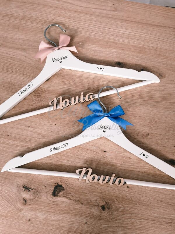 Pareja de perchas de madera personalizadas para novios en color blanco con nombres grabados Nazaret y Jesús, fechas de boda, iniciales y lazos rosa y azul de la tienda Aura's Personality